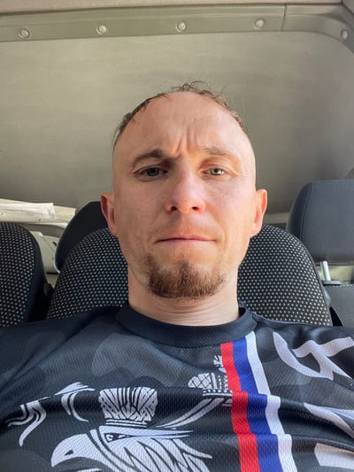 A man with a shaved head, light skin, and a goatee is seated in what appears to be a vehicle. He is wearing a black t-shirt with a graphic design featuring red, white, and blue stripes and a white crest. The man is looking directly at the camera with a neutral expression. The background shows the interior of a vehicle, including car seats and the ceiling. The lighting suggests it is daytime. There is no discernible indication of the specific location within the city of Kazan, Russia.