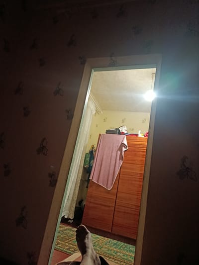 The image is a low-angle, indoor shot looking through a doorway into another room. The room visible through the doorway has a wooden wardrobe with a pink towel draped over it. The ceiling is tiled, and a light fixture is visible. The walls in both rooms are covered with floral wallpaper. In the foreground, a person's bare foot is visible, lying on a carpeted floor. The lighting is dim, with a bright light source in the second room causing lens flare. The overall impression is of a domestic setting. There is no information within the image to suggest the location is the city of Vilne, Ukraine.