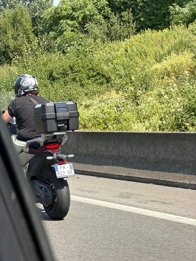 This media file shows a person riding a motorcycle on a sunny day, likely in the afternoon, given the strong shadows. The scene is outdoors, with lush green foliage and trees lining the left side of the road. The road itself appears to be paved, with white lane markings visible on the right.
The primary subject is a person on a motorcycle, viewed from behind and slightly to the side. They are wearing a black t-shirt and a helmet. The helmet is white and black with a distinct pattern and the "Arai" logo visible. The motorcycle is a modern model, predominantly black and silver, equipped with a large black storage box mounted on the rear. The license plate of the motorcycle is clearly visible and reads "FH - 927-QV". Text below the license plate appears to be in French, possibly related to the vehicle registration or manufacturer. The motorcycle also has a red taillight and a visible "HONDA" logo on the rear.
The background consists of dense green vegetation, suggesting a rural or suburban setting with ample greenery. A concrete barrier or wall separates the road from the vegetation. The overall atmosphere is bright and clear, indicative of fair weather. The image is captured from a slightly elevated perspective, possibly from another vehicle. The perspective and the presence of the rider's helmet and part of the motorcycle windshield suggest the photo was taken from a car or another motorcycle moving alongside. The location context provided is Châtenay-Malabry, France, which aligns with the presence of French license plates and typical European road infrastructure. No specific emotions are discernible from the image.