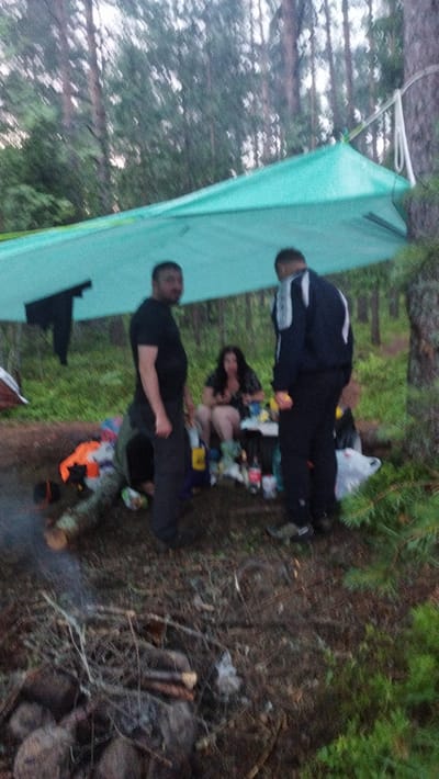 A group of four individuals is gathered in a wooded area in Tarasovskoe, Russia, under a teal-colored tarp shelter. Two men stand on the left and right of the frame, facing the center. The man on the left is wearing a black t-shirt and dark pants. The man on the right is wearing a dark jacket with white stripes and dark pants. A woman is seated in the center of the frame, wearing a black and white patterned dress. She is surrounded by various items including bottles, bags, and what appears to be food. The ground is covered with fallen leaves and branches, and a fire pit is visible in the foreground with smoking embers. The environment is a forest with tall trees and lush green foliage.