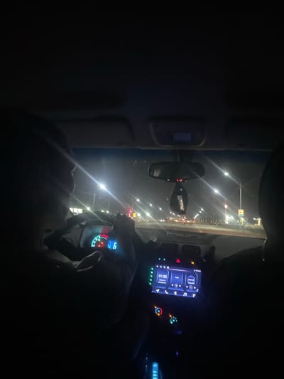 The image is taken from inside a car at night, looking forward through the windshield. Two individuals are visible, primarily their silhouettes, as they sit in the front seats. The driver is on the left, with their hands on the steering wheel, and their head is turned slightly to the right. The dashboard of the car is illuminated, showing a digital speedometer displaying "6" and a tachometer with red and blue lights. In the center console, a touchscreen infotainment system displays the time as "01:55" and a date of "2023-07-19". The view outside the car reveals a city street at night, with multiple streetlights casting bright streaks of light. There is traffic on the road, with car headlights and taillights visible. Buildings and signs are dimly illuminated in the background. A car air freshener hangs from the rearview mirror. The scene is consistent with being in the city of Ақыртөбе, Kazakhstan, given the context of the request.
