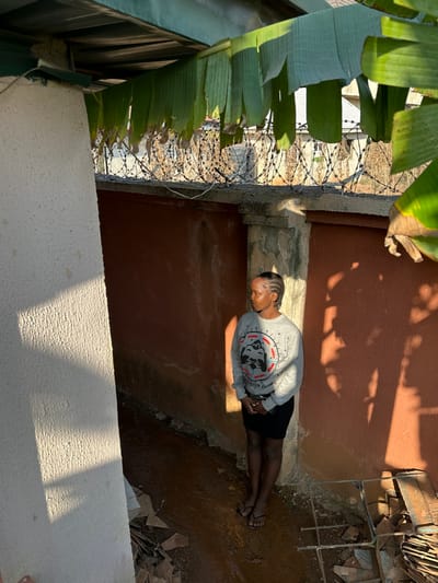 A female individual stands in an outdoor area within Agbogugu, Nigeria. The subject, with dark skin and short, braided hair, wears a grey long-sleeved sweatshirt featuring a graphic design with text, a black skirt, and black sandals. She is positioned facing slightly right, with her head turned to look towards the right out of frame, hands clasped in front. To her immediate left is a light-colored, textured wall illuminated by direct sunlight, casting shadows. Behind her is a reddish-brown wall, discolored in areas, displaying prominent shadow patterns from overhead vegetation and structures. The ground beneath is uneven, appearing wet and muddy, with scattered debris including broken tile fragments and metal scraps. Above, green banana leaves hang from a green corrugated metal roof structure. In the background, above the reddish-brown wall, a concrete perimeter wall is topped with multiple coils of barbed wire. Distant buildings and sparse vegetation are visible beyond the barbed wire under bright daylight conditions.