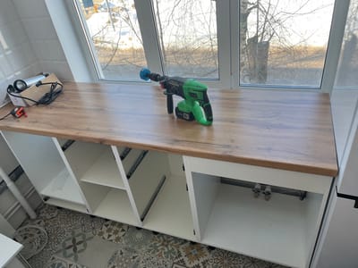 The scene depicts an interior room, likely a kitchen or utility area under construction, located in the city of Нарынка, Russia. A brown wooden-patterned laminate countertop is installed over three open, white base cabinet units. On the right side of the countertop, a green and black cordless rotary hammer drill with a blue circular attachment and a "20V" battery is positioned. To the left, a cardboard box holds a silver cylindrical object with an attached black electrical cord, beside which a small red and black tape measure rests. The base cabinets are open, revealing unfinished interiors; the leftmost unit shows a vertical grey pipe, while the rightmost unit contains two chrome water valves and visible metal drawer slides. The floor is tiled with ceramic tiles featuring a varied geometric and floral pattern in shades of grey, beige, and blue. A large white-framed window spans the background, looking out onto an exterior scene with bare tree branches and patches of snow on the ground under natural light, indicating a colder season. A white tiled wall is partially visible on the far left. No individuals are present in the image.