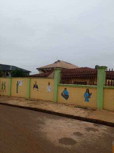 The image shows a yellow wall with green trim, painted with various figures of people, in front of a building, at coordinates 7.294, 5.163, in the city of Ita Oniyan, Nigeria. The wall is adjacent to a paved street. On the wall, there are five figures painted in various colors: two are of men wearing white shirts and dark trousers, one is of a man wearing a blue suit and dark trousers, one is of a man wearing a blue shirt and dark trousers sitting at a desk, and one is of a woman wearing a blue dress and a brown hat, holding a book. The building behind the wall has a brown roof with faded, brown tiles. A brown, wooden fence, and a black fence post are located on the left side of the wall. Another black fence post is visible on the right side of the wall. There is a cloudy, gray sky.