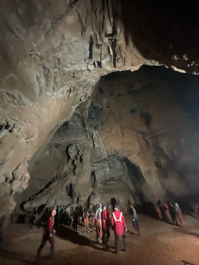 The image shows a group of people inside a large cave. The cave walls are rough and textured, with visible rock formations and shadows that suggest a cavernous space. The lighting is dim, with some areas illuminated by artificial light sources, casting long shadows. The people are dispersed throughout the cave, some standing in groups, others walking. They are dressed in casual clothing, with a few individuals wearing brightly colored vests. The ground is covered with dirt and rocks. The overall scene suggests an exploration or tour of a natural cave formation. The location is identified as the city of Gollagutta, India, a region known for its numerous cave systems.
