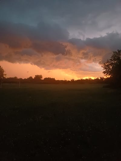 This image captures a dramatic sky over a field, likely at sunset or sunrise, given the intense orange and pink hues illuminating the clouds. The sky on the left is dominated by dark, brooding clouds that transition into a vibrant band of sunset colors towards the horizon. Silhouetted trees form a dark line along the edge of the field, with hints of leaves visible against the bright sky.
The field itself appears to be a grassy area, possibly a park or sports field, with a soccer goal visible in the mid-ground. The grass is dark, suggesting it is either evening or the lighting conditions are low. A few scattered lights or stars are visible in the dark areas of the image, potentially indicating the presence of fireflies or distant lights.
The overall mood is one of natural beauty and awe, with the juxtaposition of the dark, stormy clouds and the radiant sunset. The scene evokes a sense of the transition from day to night, with a powerful display of atmospheric phenomena. The location is identified as Trypillia, Ukraine, which adds a geographical context to the otherwise universal depiction of a striking sky. There is no visible text in the image.
