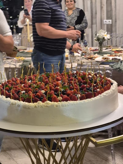 The image depicts a celebratory event with a large, round cake as the central focus. The cake is covered in white frosting and topped with an abundance of strawberries, blueberries, and small green leaves. Several thin, gold skewers are stuck into the fruit, creating a decorative effect. In the background, people are visible, suggesting a gathering or party. One individual is wearing a striped shirt and blue jeans, with their arm extended as if reaching for something. Another person in the background is wearing a black and silver patterned top and is smiling. The setting appears to be indoors, with a table laden with food and drinks in the background. The lighting suggests an evening event. The location is identified as Zelyonovka, Russia.