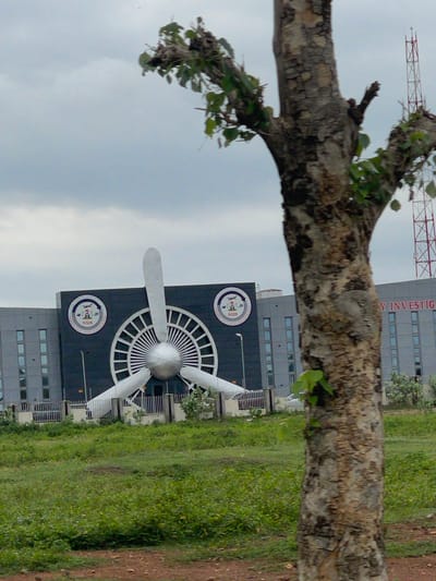In Immigration Quarters, Nigeria, the image captures the Nigerian Safety Investigation Bureau (NSIB) headquarters, an imposing building with a prominent propeller sculpture on its facade. The propeller, rendered in a metallic silver, features three blades and a central spherical hub, set against a dark background adorned with circular emblems depicting an airplane and the text "NSIB" and "NIGERIAN SAFETY INVESTIGATION BUREAU". The building itself is modern, with a repeating pattern of grey rectangular windows across its grey exterior.
In the foreground, a large tree with textured bark and sparse green foliage partially obstructs the view of the building. The tree's branches extend towards the top of the frame, with some leaves appearing to be in the process of shedding. Behind the building, a tall red and white lattice tower, likely a communication or broadcasting antenna, is visible against an overcast sky. The ground in front of the building is covered with a low, green grassy area and a red dirt path in the immediate foreground, suggesting an outdoor, possibly semi-rural or developing urban setting. No people are visible in the image. The overall scene is static and focused on the architectural features of the NSIB headquarters and its immediate surroundings.