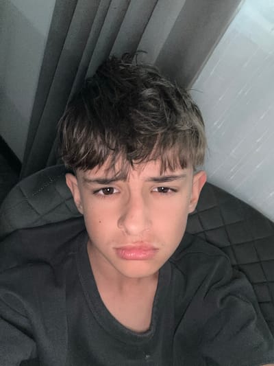 A young person with light skin, brown eyes, and short, highlighted brown hair is lying down, resting their head on a quilted dark grey or black cushion or surface. The person is wearing a dark grey or black t-shirt. The background to the left is a grey textured surface, possibly a wall or another piece of furniture. The overall lighting is somewhat dim. There is no indication of the location being Slokoshtitsa, Bulgaria or any city. There are no actions or interactions. The scene is a single person posed for a photo.