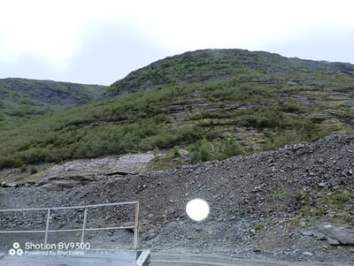 The image depicts a rocky, overgrown mountainside in Koashva, Russia. The foreground features a gravelly slope leading up to the mountain, with a metal railing on the left side. A bright, circular light source, possibly a reflection or lens flare, is visible on the gravel. The mountainside itself is covered in dense green vegetation and interspersed with rocky outcrops. The sky above is overcast and hazy. The lower left corner of the image contains a watermark indicating it was "Shot on BV9300 Powered by Blackview".