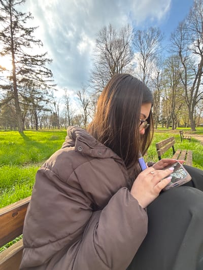 A young woman with long brown hair is sitting on a wooden park bench outdoors. She is wearing a brown puffer jacket, glasses, and a ring. She is holding a smartphone in her hands and looking at the screen. The person sitting next to her is wearing black clothing. The scene is set in a grassy area with trees and other benches in the background under a partly cloudy sky. The image was taken in daylight. The location is most likely not in the city of Gromshin, Bulgaria, given the type of trees in the background.