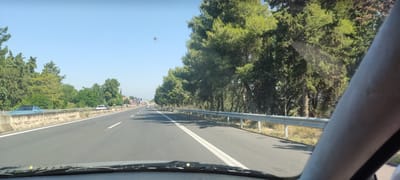 The image presents a forward-facing view from inside a vehicle traveling on a multi-lane asphalt highway in Kardia, Greece, under clear blue sky conditions. The vehicle's dashboard and a portion of a windshield wiper are visible at the bottom of the frame. On the far right, a segment of a human arm and the vehicle's A-pillar are present.

The highway features clearly defined white lane markings, including a solid line delineating the shoulder on the right and dashed lines separating traffic lanes. A metal guardrail runs along the right side of the road, bordering a verge with dry vegetation and a dense line of tall green conifer-like trees.

On the left side of the highway, opposing traffic lanes are separated by a concrete barrier topped with a metal guardrail. Beyond this barrier, a variety of green trees and foliage are visible. In the opposing lanes, a light blue convertible car is parked or stopped behind the barrier, followed by a white pickup truck and another dark vehicle further down the road. In the far distance on the left, several structures, including buildings and what appears to be commercial signage, are discernible. Multiple vehicles are observed in the distance on the same side of the highway, indicating traffic.

A prominent reddish-brown spherical artifact, consistent with sensor dust or a lens anomaly, is visible in the upper-center portion of the sky. The scene is illuminated by bright daylight.