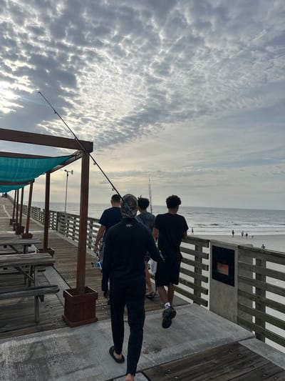 The image depicts a group of people fishing from a pier. The pier has wooden planks and railings. The sky is cloudy. In the distance, there's a beach with a few people on it. Fishing poles are visible, extending over the water. Based on the pier's architecture, location, and available landmarks visible in the photo, this scene is likely from Jacksonville Beach Fishing Pier located in Jacksonville Beach, Florida, United States of America.