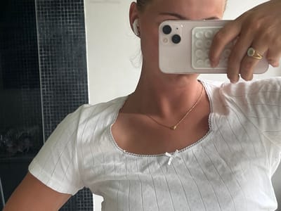 A partial view of a person is captured in a mirror selfie within an indoor environment. The subject, a young adult female, is visible from the upper torso to just below the neck, with blonde hair partially seen on the right. She wears a white, short-sleeved top with vertical ridging, subtle perforations, a square neckline edged with white scalloped lace, and a small white bow centered at the bottom of the neckline. A thin gold-colored necklace with a small heart-shaped pendant rests on her collarbone. Her right ear features a white wireless earbud. She holds a light-colored smartphone, likely an iPhone, in her right hand, obscuring most of her face. The phone's rear dual cameras are visible, along with a white PopSockets-style grip attached to its back. A gold-colored ring with a circular, light-colored stone set in a broad bezel and double band is on her right ring finger. The background consists of a dark, glossy, small-tiled wall on the left, displaying a faint, dark reflection of an indeterminate object or domestic animal, and a plain white wall on the right. The specific location in Skanderborg, Denmark, is not discernible from the visual information presented.
