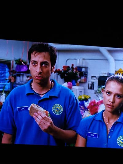 The image is a medium shot of two people, a man and a woman, wearing blue polo shirts with "AQUA WORLD STAFF" patches. The man, on the left, has dark hair and is holding a sandwich. A name tag on his shirt reads "JOE". The woman, on the right, has blonde hair pulled back and is wearing a necklace. A name tag on her shirt reads "CAM". They appear to be in an indoor setting with shelves and floral arrangements in the background. The scene is illuminated by bright, even lighting. This image is not in the city of Turka, Ukraine.