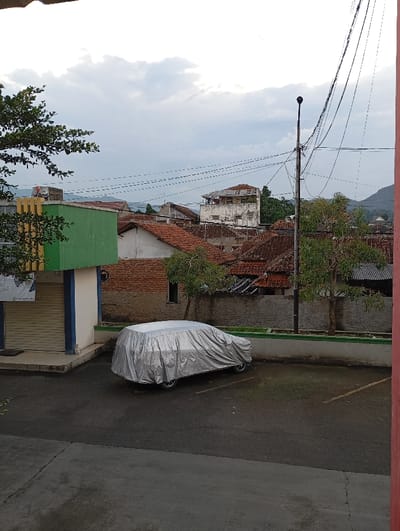 The image displays an urban scene in Pamekasan, Indonesia, under an overcast sky. In the immediate foreground, a dark asphalt parking area is visible. Centrally positioned on the asphalt is a vehicle entirely covered by a silver protective tarp. To the left, a section of a single-story building with light green walls and blue trim features a closed metallic roller shutter at its entrance, with a small dark mat on the ground outside. A low wall with a green coping separates the parking area from the background.

Beyond this wall, a dense cluster of residential buildings with reddish-brown tiled roofs extends across the midground. A distinct, multi-story white building, appearing weathered with a damaged roof structure, is visible towards the right-center of this cluster. A tall black utility pole, laden with numerous intertwined overhead power lines, stands prominently on the right side of the midground. Sparse trees with green foliage are dispersed among the buildings and behind the dividing wall. In the distant background, faint mountain ranges are discernible beneath the gray, cloudy sky. No individuals are present in the captured scene.