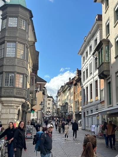 The image depicts a street scene in Schaffhausen, Switzerland, characterized by narrow cobblestone streets lined with historic buildings. The buildings showcase a mix of architectural styles with ornate facades, multiple windows, and varied rooflines. A bright blue sky with a few clouds is visible above.

The street is populated with people walking in both directions, appearing as tourists or locals. A couple of people are standing near a Jack & Jones store, as well as a sign advertising "DROGISTORE". There are visible shops on either side of the street. The photo is taken at street level.