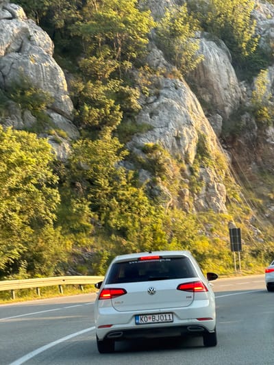 The image shows a white Volkswagen Golf TDi driving on a paved road. The car has a license plate that reads "KO BJ011" and a sticker on the rear window with the text "www.oasispmontenegro.me". The car is in motion, suggested by the blur of the surroundings and the car itself.

The road appears to be a scenic route, possibly a highway, flanked by a steep, rocky hillside covered in dense green vegetation. The sun is shining brightly, casting long shadows and illuminating the foliage with a warm, golden light. The sky is not visible in the frame. A metal guardrail runs along the side of the road.

In the background, another white car is partially visible, also in motion. A dark, rectangular traffic sign or utility box is positioned on the side of the road further up. The scene is likely taking place in a mountainous or hilly region. Based on the license plate and the website sticker, the location is suggested to be Markovići, Montenegro.