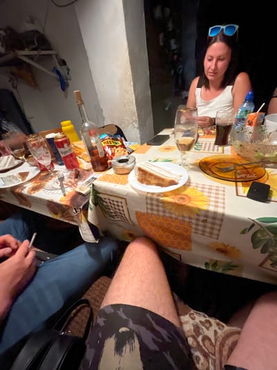 The image depicts a dimly lit outdoor gathering, likely at night, possibly in the backyard or a common outdoor space of a residential property in Dragoevo, Bulgaria. A group of people are seated around a table covered with a tablecloth featuring sunflowers and a checkered pattern. The table is laden with food and drinks, including glasses of amber-colored liquid, a bottle of alcohol, a can of soda, a bag of snacks, and slices of cake on plates. A woman in a white tank top, with blue sunglasses perched on her head, is seated at the table. Another individual is holding a lit cigarette. The environment appears somewhat cluttered, with shelves and objects visible in the background, suggesting a casual and informal setting. The overall atmosphere is one of a relaxed social gathering.