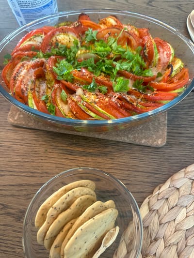 The image displays an overhead view of a dining scene set on a light brown wooden table. The primary subject is a clear, oval-shaped glass baking dish containing a cooked vegetable gratin. The dish features thinly sliced red tomatoes and green zucchini, meticulously arranged in overlapping concentric layers. The vegetables exhibit some browning on their edges, indicative of baking, and are topped with a sparse scattering of grated cheese and fresh green parsley sprigs. A reddish-orange spice is also visible on the surface. This dish rests on a rectangular, light brown trivet with a rough, textured surface.
In the lower left foreground, a small, clear glass bowl holds several light beige, oval-shaped crackers containing dark seeds. To the right of the cracker bowl, a portion of a woven placemat, crafted from light brown natural fibers, is visible. In the upper left background, a white plastic container, partially obscured and oriented sideways, displays Portuguese text including "QUEIJO FETA D.O.P." and "FETA F.O.B. 43%." No individuals are present in the image, and no specific actions or interactions are depicted beyond the presentation of the prepared food items. The environment is an indoor setting. There are no visual cues within the image that allow for the specific identification of the location as Ericeira, Portugal, although the Portuguese text on the container is consistent with the country.