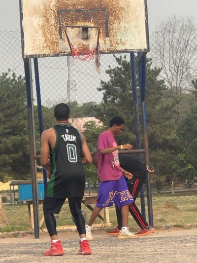 The image depicts an outdoor basketball court in Umunekwu, Nigeria, featuring three individuals and a basketball setup. A heavily rusted, white-colored rectangular basketball backboard with blue support poles stands centrally, fitted with a hoop displaying an incomplete net composed of red, white, and blue segments.
In the foreground, a person is seen from the back, wearing a black and green sleeveless basketball jersey with "TATUM 0" printed on it, black compression leggings, and red high-top athletic shoes. To their right, a second individual wears a magenta t-shirt, purple shorts with "LAKERS" written on them, a yellow wristband on their left arm, and light-colored athletic shoes. This individual is looking downwards, with hands positioned forward. A third person is partially visible behind the second individual, bent over at the waist, wearing black clothing with red stripes on the pants and orange and blue athletic shoes.
The ground surface appears to be a light-colored concrete or asphalt court. A chain-link fence runs horizontally across the mid-ground, separating the court from the background. Beyond the fence, various trees with green foliage are visible, along with a distant lattice-style communication tower and a partial view of a light-colored building. The sky is overcast.