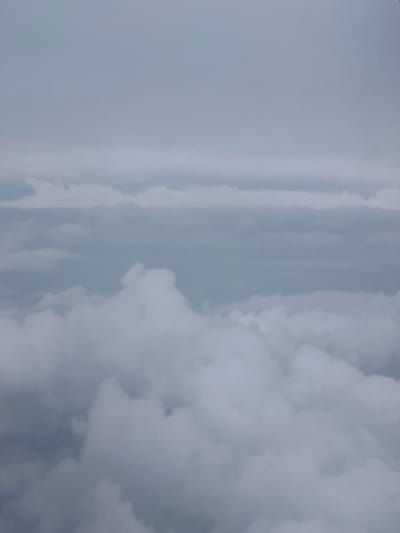 The image, oriented vertically, captures a sky heavily populated with various cloud formations, possibly seen from an aerial perspective above the Spata-Loutsa Municipal Unit, Greece. The predominant colors are shades of grey and white, indicative of an overcast or partially obscured sky during daylight.
On the left side of the frame, the sky transitions from a lighter, more uniform grey at the extreme left to areas with subtle, elongated cloud streaks. As the view progresses towards the right, the cloud cover becomes significantly denser and more textured. The right side of the image features voluminous, cumulus-like clouds with distinct peaks and valleys, showcasing deep shadows in their recesses and brighter highlights on their surfaces, suggesting considerable depth and mass.
No human figures, man-made objects, or visible text are present in the image. The scene conveys a sense of vastness and atmospheric layering, typical of cloudy weather.