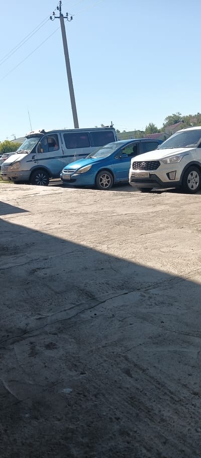 The image depicts a street scene in Okoneshnikovo, Russia, on a sunny day with a clear blue sky. In the foreground, a cracked concrete surface is visible, with a large shadow cast across it from an unseen object. Behind the concrete, several vehicles are parked. From left to right, there is a silver van, a blue hatchback, and a white SUV. A utility pole with wires extends upwards in the background. The vehicles appear to be parked on asphalt. The overall environment suggests an outdoor parking area in a town or city. There are no people clearly visible in the image.