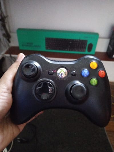 This image, captured in Villanueva de Gállego, Spain, depicts an indoor scene focused on a black Xbox 360 wireless controller. A human hand, with a light to medium skin tone, grips the left side of the controller, with the thumb and part of the palm visible. The controller is oriented with its face buttons visible: a black left analog stick (showing minor wear), a black D-pad, a reflective silver Xbox guide button, black 'Back' and 'Start' buttons, and four colored action buttons – yellow 'Y', blue 'X', red 'B', and green 'A'. A black right analog stick is also visible. The controller's surface exhibits minor scuffs and dust.
In the background, a dark, textured surface is visible directly beneath the controller. Above this, a horizontal wooden surface extends across the frame. Resting on the wooden surface is a rectangular, out-of-focus green object, which appears to have a dark, grid-like pattern resembling a keyboard. The uppermost background consists of a plain, light-colored wall. The scene is illuminated by ambient indoor light.