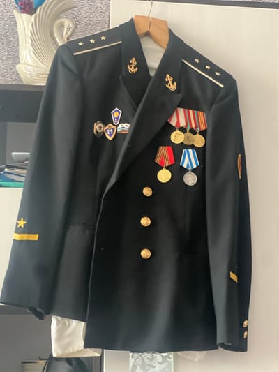 The image shows a dark navy blue formal jacket, likely a military or naval uniform, hanging on a wooden hanger. The jacket is adorned with several medals and pins. On the left shoulder, there are four gold stars on a dark band with a white stripe, and an anchor pin. On the right shoulder, there is an anchor pin. Across the chest, there are several rows of medals with colorful ribbons and circular medallions. There are also gold buttons on the front of the jacket. On the left sleeve cuff, there is a yellow chevron with a yellow star above it. On the right sleeve, there is a yellow chevron. A white shirt is visible underneath the jacket. The background shows a shelf with a decorative white swan figurine and some papers. The lighting suggests an indoor environment, possibly a home. The image does not provide any specific information about the location in the city of Sherbakul', Russia.