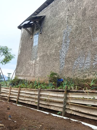 A weathered, textured building stands beside a garden enclosed by a low fence constructed from horizontal slats. The fence is supported by vertical posts, and the garden contains leafy plants. A small window with a rudimentary overhang is visible on the building. The ground is muddy and appears recently tilled. Part of mountains and sky peek from the left side.