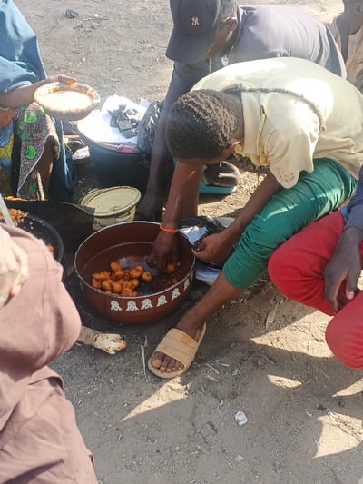 The image portrays an outdoor scene in Gobirawa, Nigeria, featuring multiple individuals gathered around food items on unpaved dirt ground under bright sunlight. In the central foreground, a person with short, dark hair, wearing a light yellow short-sleeved shirt, green trousers, and beige sandals on their left foot, squats. This individual's hands are inside a large brown bowl with a white decorative pattern around its rim, actively handling numerous small, round, golden-brown fried dough balls and some darker, smaller items, possibly beans or seeds. An orange wristband is visible on their left wrist, and a dark beaded necklace hangs around their neck. They also interact with a black plastic bag or container held in their right hand.

To the immediate left of the central person, a black cooking pot or pan containing similar fried items is visible, along with a yellow enamel pot with a lid resting on the ground. Further left, a partially obscured individual, dressed in a blue garment over a colorful patterned fabric, holds a metal bowl containing a light, creamy substance. Behind the central figure, another individual, wearing a dark baseball cap with a white "NY" logo and a dark grey shirt, is partially visible, leaning over the scene. To the far right, the lower body of a person wearing red trousers is visible. The ground is characterized by light brown, dry earth with scattered small debris and prominent shadows cast by the subjects, indicating strong overhead sunlight. In the background, a black plastic basin contains other items, including dark, possibly dried, food pieces wrapped in paper or plastic.