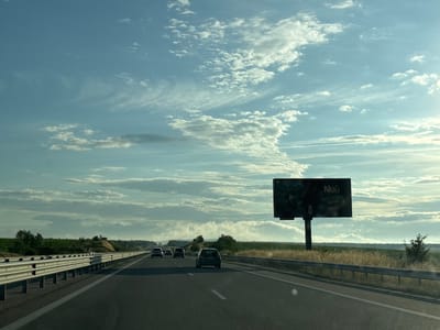 A wide-angle, eye-level shot captures a highway scene under a partly cloudy sky with the sun shining through. The highway is divided by a metal guardrail, with two lanes visible in the foreground traveling away from the camera. Several cars are driving on the highway, including a dark-colored hatchback in the right lane in the foreground. To the left of the highway, another metal guardrail runs parallel to the road, separating it from a grassy verge and a field with some vegetation and trees in the distance. A large billboard stands to the right of the highway, featuring text that appears to read "Null". The sky is filled with wispy, elongated clouds against a light blue backdrop. The overall impression is of a travel or scenic view along a Bulgarian highway, possibly near the city of Malko Tarnovo.
