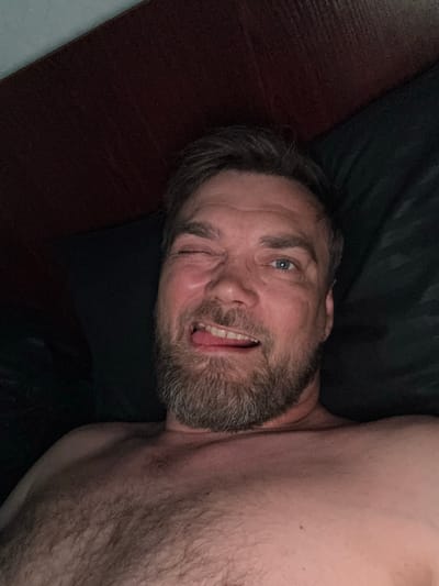 A selfie of a man in his late 30s or early 40s, lying down. He has short brown hair, blue eyes, and a beard. He is winking and sticking his tongue out slightly, with a playful expression. The man is shirtless, and his chest and shoulder are visible. He appears to be indoors, possibly in bed, as suggested by the dark, soft bedding in the background. The lighting is dim, suggesting it might be nighttime or a dimly lit room. The image is taken from a high angle, looking down at the man. There is no visible text in the image.