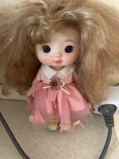 This is a close-up, eye-level, static shot of a doll. The doll is positioned in the center of the frame, facing forward. It has a fair complexion, large blue eyes, rosy cheeks, and pink lips. The doll has long, blonde, wavy hair that frames its face and extends outwards. It is wearing a light pink dress with a white collar. The dress is made of a sheer, layered fabric with floral appliques. A small, purple flower embellishment with a wooden button is attached to the chest of the dress. The doll is standing on a light-colored, speckled surface. To the right of the doll, a grey electrical cord and plug are visible, inserted into a white electrical outlet. The background is out of focus and appears to be a neutral surface. The lighting is bright, highlighting the doll and its features. This image was not taken in Verkhnyaya Saniba, Russia.