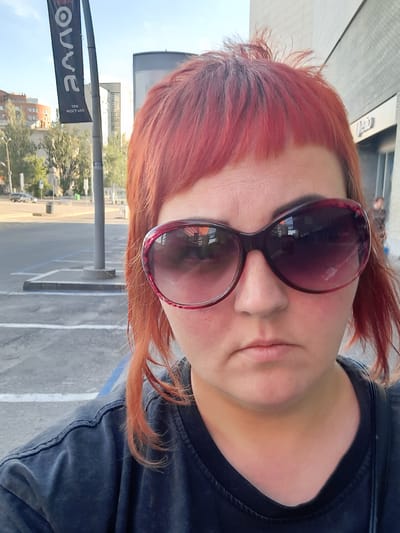 A medium shot shows a woman with bright red hair and large, dark sunglasses. She is wearing a dark t-shirt. The woman is outdoors, and in the background, there is a parking lot with white and blue markings, a lamppost, trees, and buildings. A banner on the lamppost has "OLVE" written vertically in large white letters. The building behind her appears to be modern, with a grey facade and windows. The scene is consistent with an urban environment.