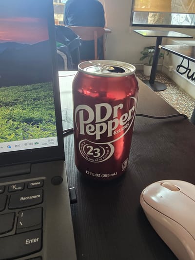 A can of Dr. Pepper sits on a dark wooden table next to an open laptop, a pink computer mouse, and a black USB receiver. The laptop screen displays a vibrant landscape of rolling green hills under a sunset sky. The Dr. Pepper can is partially opened, with the lid ajar. In the background, through a window, a small potted plant sits on the floor beside a black table. The scene suggests a casual work or leisure environment. The image location is the city of Klapanunggal, Indonesia.