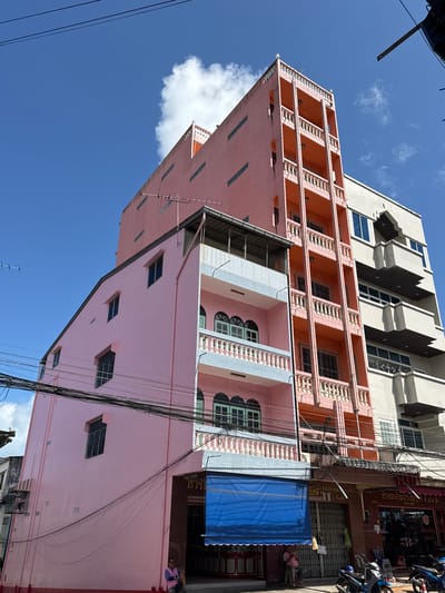 This image depicts a street scene in Ranong, Thailand, featuring a row of multi-story buildings. The primary building on the left is a narrow, tall structure painted a bright pink, with a sloped roofline and several windows. Adjacent to it is a taller, salmon-pink building with a prominent vertical section featuring multiple balconies with white balustrades and arched window openings. To the right of this is a lighter-colored building, possibly cream or beige, with a more modern facade and a series of horizontal dark-colored overhangs or balconies. The buildings are situated along a street with overhead power lines crisscrossing the frame. A blue awning is visible on the ground floor of the pink building. In the foreground, several motorcycles are parked. Two individuals are partially visible: one sitting on a ledge in front of a storefront on the ground floor of the pink building, and another person near the motorcycles. The sky is a clear, vibrant blue with a few scattered white clouds. The architecture suggests a densely populated urban environment, likely a commercial or mixed-use area.