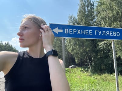 A young woman stands outdoors in Verkhnee Gulyaevo, Russia, a rural setting with a clear blue sky and lush green trees. She is looking to her left, with her right hand touching her hair. She is wearing a black tank top, a smartwatch on her left wrist, and white wireless earbuds. A blue sign pointing to the left with white Russian text reading "ВЕРХНЕЕ ГУЛЯЕВО" (Verkhnee Gulyaevo) is visible to her right. In the background, a light-colored car is parked on a grassy slope, surrounded by trees. The overall scene is bright and sunny.