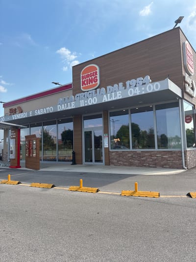The image shows a Burger King restaurant in Paese, Italy. The restaurant is a modern building with large windows and a drive-thru. The Burger King logo is prominently displayed on the building. There is also a sign with the text "ALLA GRIGLIA DAL 1954 VENERDI E SABATO DALLE 11:00 ALLE 04:00". The weather is sunny and the sky is blue. There are no people visible in the image. The scene is a typical fast-food restaurant setting.