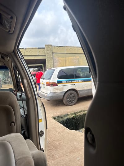 The image captures a street scene in Orji, Nigeria, as viewed from inside a vehicle. In the foreground, the interior of a car is visible, with beige-colored seats and a portion of the dashboard. Outside the car, a white van with blue and yellow stripes is parked. The van has the text "ENUGU STATE TRANSPORT CO. LTD." printed on its side. A person wearing a red shirt and blue pants is standing next to the van, facing away from the camera and looking towards the left. Behind the van, a concrete bridge or overpass is visible, with what appears to be a market stall or shops in the background. The ground is dusty and uneven, with a drainage ditch running alongside the road. The sky is overcast, suggesting a cloudy day.