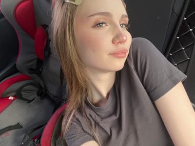 A young Caucasian female with blonde hair, light skin, and green eyes is seen in a close-up, side-profile shot. She is wearing a grey t-shirt and has on makeup, including mascara and lip gloss. Two light-colored hair clips are visible in her hair. She appears to be sitting in a red and grey car seat, indicated by the textured fabric and red cushioning visible in the background. The environment suggests she is in or near a vehicle, likely a car. The location is not discernible from the image itself; it is not possible to determine if the image was taken in Новая Отрадовка, Russia.