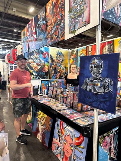 A man and a woman are at an art exhibition in Villepinte, France. The man, wearing a red t-shirt and camouflage shorts, stands by a table displaying various anime-style art prints. The woman, in a black off-the-shoulder top, stands next to a large painting of a robot. The exhibition appears to be an indoor event, possibly a convention, with the artwork displayed on walls and tables. The overall atmosphere is vibrant and colorful, with many detailed illustrations featuring anime characters and themes.
