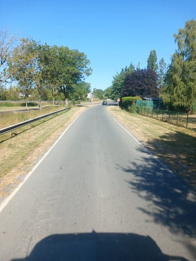 A road curving to the left with a guardrail on the left side of the image. Trees and vegetation line both sides of the road, with a dark green structure visible on the right. A black car is driving away from the camera on the left side of the road, and a traffic sign indicating a right turn is visible near the car. The sky is clear and blue. The image appears to be taken from a moving vehicle, as indicated by the shadow of the vehicle on the road. The location is Péronne, France.