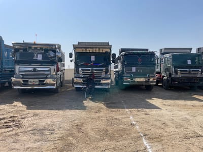 The image displays an outdoor scene in Pir Dust, Afghanistan, featuring a row of five large dump trucks parked on a dry, unpaved, light brown dirt lot under a clear, cloudless blue sky. A single individual, clad in dark attire, is seated on a black folding chair positioned between the second and third trucks from the left. The trucks are primarily Hino brand, with four distinctly visible Hino logos on their grilles. All trucks possess dump bodies, and several exhibit custom detailing on their cabs, including chrome grilles, additional lighting, and decorative elements such as calligraphy and small flags on the windshields and roofs. White rectangular papers are affixed to the windshields of at least four of the trucks. A faint white line is painted horizontally on the dirt ground in the foreground. In the background, a light-colored wall topped with wire fencing runs horizontally behind the vehicles.