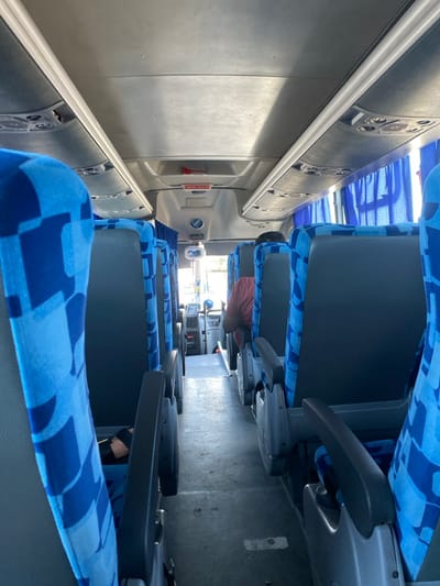 Inside a bus, rows of seats upholstered in a blue geometric pattern line the aisle. Overhead compartments run along one side, featuring speakers and a red emergency handle. Passengers are seated, some visible in partial view between the seats. The floor is gray and worn. Light streams in from the windows.
