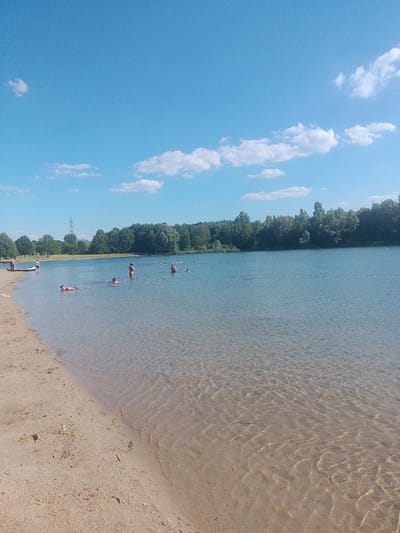 This is a full-shot, eye-level, vertical media file of a sunny day at a lake in Lünen, Germany. The scene depicts a sandy beach on the left, transitioning into clear, shallow water, with a treeline bordering the far side of the lake. The sky is a vibrant blue with scattered white clouds.
Several people are visible in the water and on the beach. In the shallow water near the beach, a man is lying on his back, seemingly relaxed. Further out, other individuals are swimming or wading. On the beach, a few people are visible near a small boat or raft.
The overall atmosphere is one of a peaceful summer day at a recreational lake. The sunlight glints off the water, creating ripples and reflections on the sandy lakebed, indicating the shallow depth near the shore. The trees in the background are lush and green, suggesting it is summertime. The clear sky and bright sunlight point to fair weather and likely daytime, possibly mid-afternoon. There is no visible text in the media file.