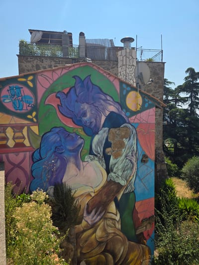 A mural depicts two figures, one with blue skin and purple hair, leaning into the embrace of another figure with purple, wild hair and facial features resembling a beast. They are set against a colorful background of geometric shapes and floral motifs. The mural is painted on the side of a building, with portions of the wall exposed. Above the mural, the rooftop of the building is visible, featuring a satellite dish, a chimney, and a covered area. Trees and vegetation surround the building. The mural is located in the historic center of Viterbo, Italy, specifically on the wall of Palazzo degli Scudi.