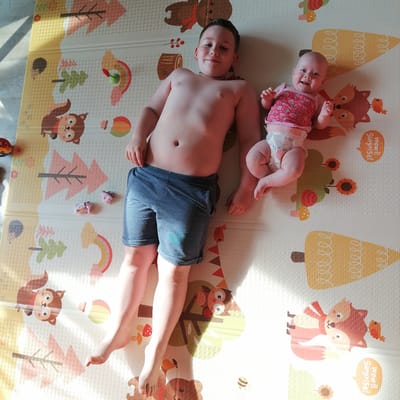 This is a top-down, eye-level photograph of two young children lying on a soft, padded play mat. The mat features a whimsical woodland theme with cartoon illustrations of animals like squirrels, foxes, bears, and hedgehogs, as well as natural elements such as trees, leaves, mushrooms, and a rainbow. The lighting suggests it is daytime, with sunlight casting shadows across the mat, indicating a sunny environment.
On the left, a young boy, likely around 6-8 years old, is lying on his back. He is shirtless and wearing blue denim shorts. His expression is a slight smile, suggesting contentment or amusement. Next to him, on the right, a baby girl is lying on her back. She is wearing a pink, strawberry-patterned sleeveless top and a diaper. She is smiling widely and looking upwards, her tiny hands outstretched. The baby appears to be around 3-6 months old.
Several small objects are scattered on the mat near the boy, including three pairs of children's socks, a small toy car, and a small red and white mushroom toy. The overall scene conveys a sense of a relaxed and happy moment at home, likely in a nursery or play area. There is no visible text in the image, other than small decorative text on the play mat that is illegible. The location context of Trubacheyevka, Russia, is provided but not visually discernible from the image itself.