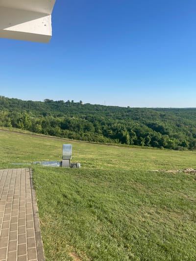 The image depicts a scenic outdoor setting in Stăuceni, Moldova, on a clear, sunny day. The scene is dominated by a vibrant blue sky and a vast expanse of lush green grass. In the foreground, a modern concrete structure with sharp angles is partially visible on the left side.
To the right of the grassy field, a paved pathway made of light-colored bricks runs along the edge. Further into the field, there are some metallic structures that appear to be ventilation units or equipment, one of which is a prominent silver rectangular box. A dirt path winds through the grassy area, leading towards a densely forested hillside that rises in the background. The trees are a rich green, suggesting a temperate climate and likely summer or late spring.
The overall impression is one of a well-maintained outdoor space, possibly a park, recreational area, or the grounds of a facility. The bright sunlight indicates it is daytime, likely mid-morning or afternoon. There are no people visible in the image. The focus is on the natural landscape and the man-made elements within it. The composition, with the sky occupying a significant portion of the frame, emphasizes the open and airy atmosphere.