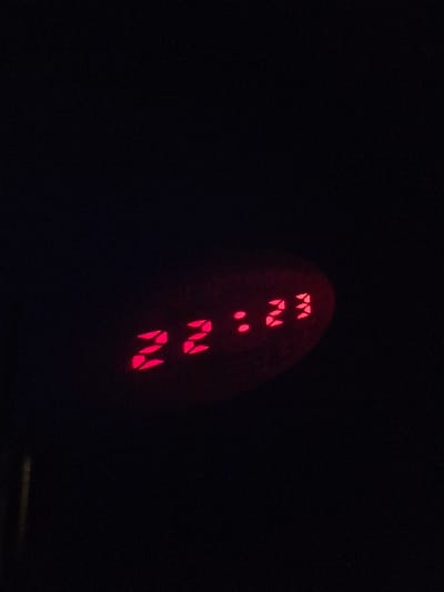 The scene depicts a digital clock displaying "22:23" in a dark, low-light environment. The time is rendered in red, segmented LED-style numbers, partially enclosed by a faint, dark elliptical or oval outline. A thin, vertical, light-colored reflection or object edge is visible along the far left margin of the image. No people, specific objects beyond the clock display, or discernible actions are present. The location is specified as the city of Niedernhausen, Germany.